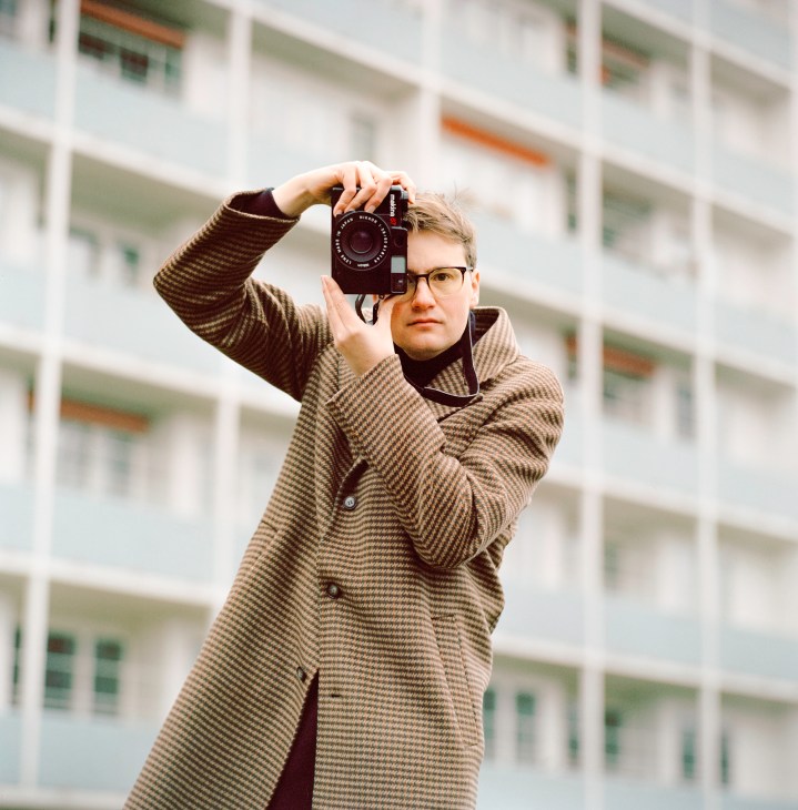 Robert Rieger holding a vintage camera up to his eye while wearing glasses and a brown checkered coat