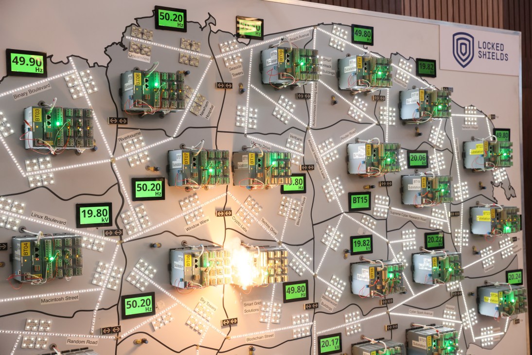 Power grid control system display showing circuit boards, monitoring equipment, and frequency readings on green digital displays.