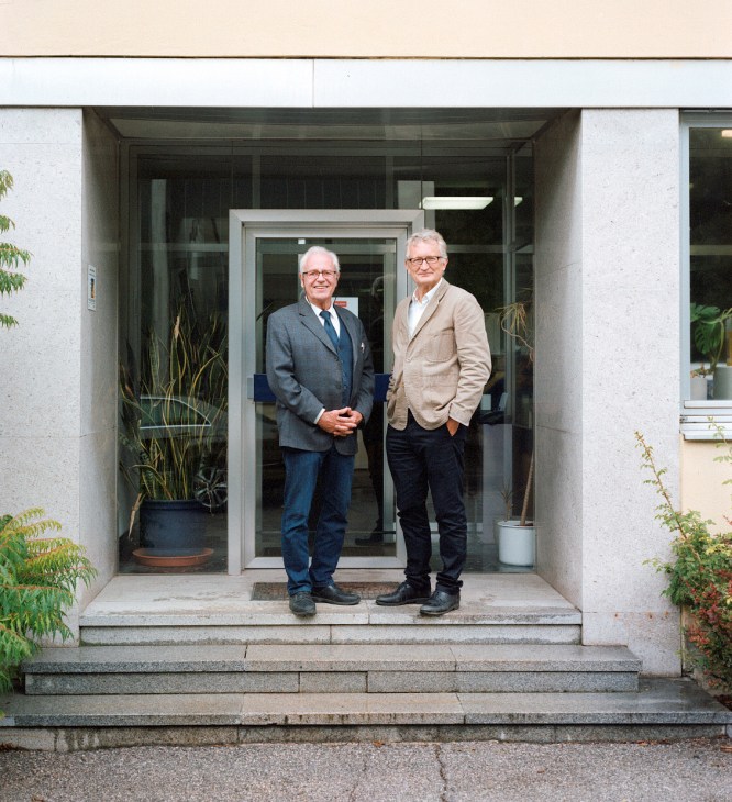 Two men stand in the doorway of a modern minimalist house with glass doors and white walls.