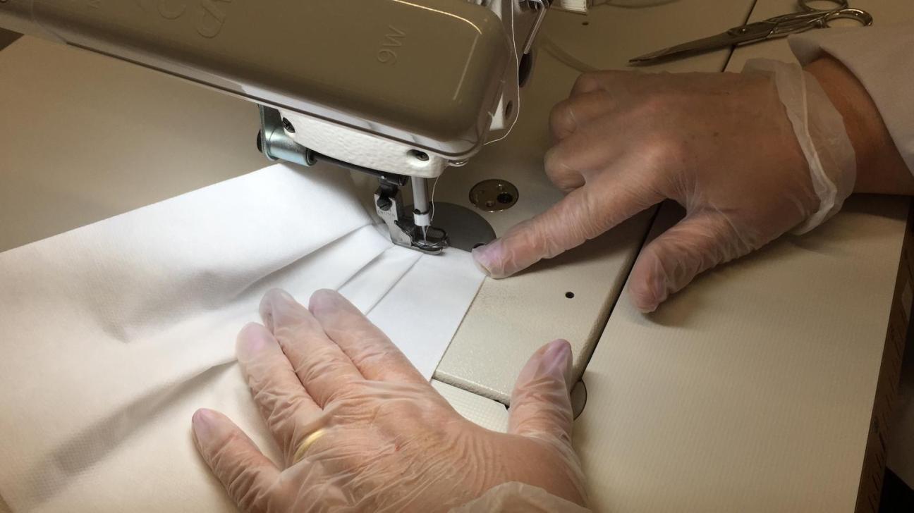 Hands operating a sewing machine stitching white fabric for mask production.