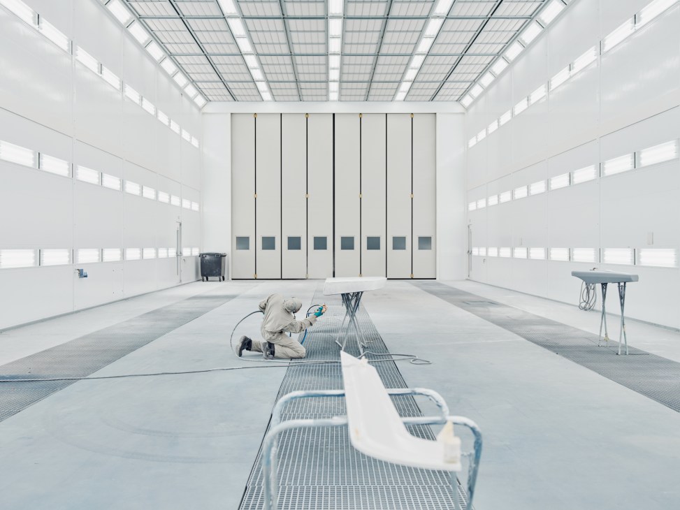 Worker spray painting white boat hull sections in a bright, modern industrial facility with solar panels.