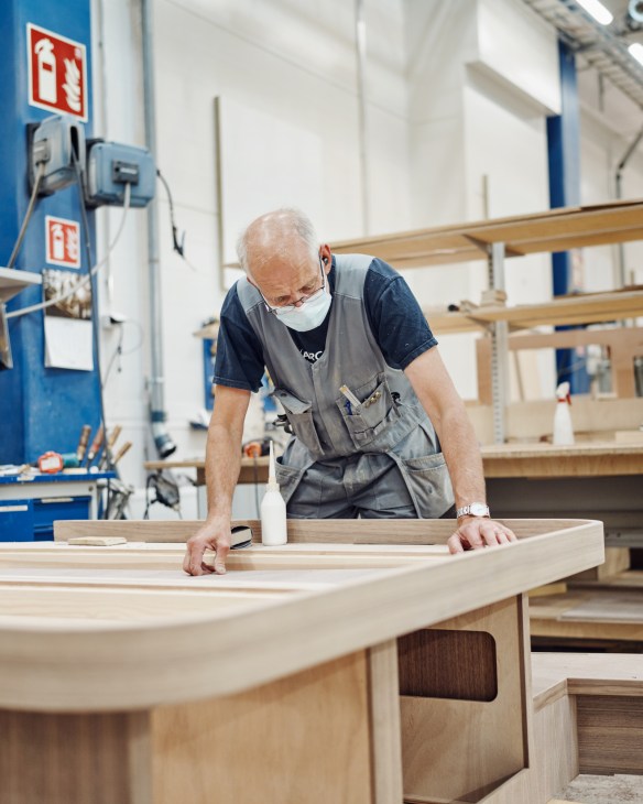 Craftsman in apron and face mask sanding curved wooden boat hull at Nautor yacht factory workbench.