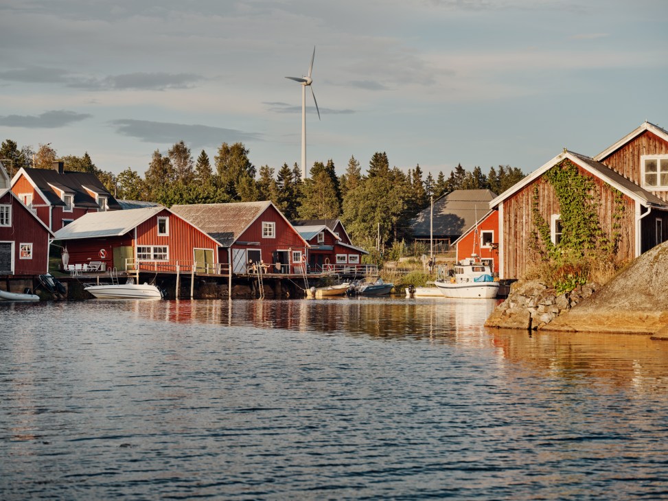 Red wooden fishing huts on stilts by the water in Skeppsmalen, Sweden, with coniferous forest and a wind turbine behind.