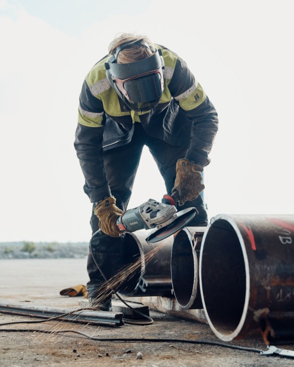 Worker in safety gear welding a large metal pipe at a port with water visible in the background.