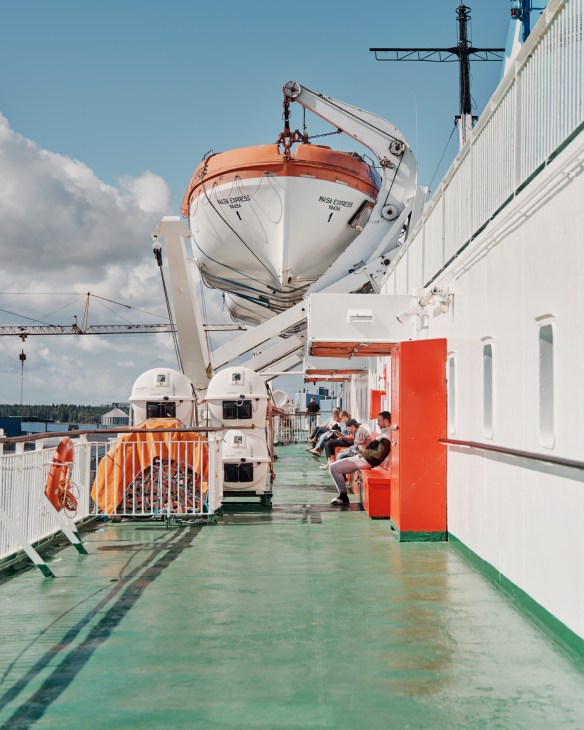 Passengers sitting on the deck of the Wasa Line ferry with a lifeboat suspended above.