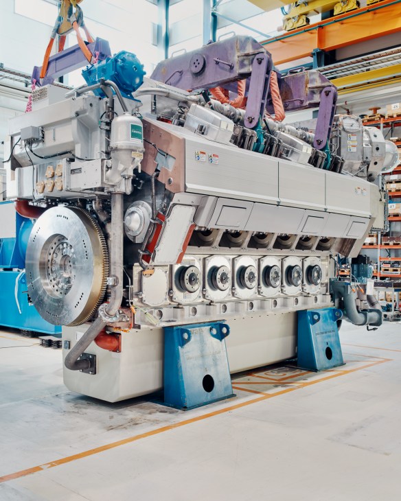 Large marine engine with blue and purple components suspended by overhead crane at Wärtsilä factory.