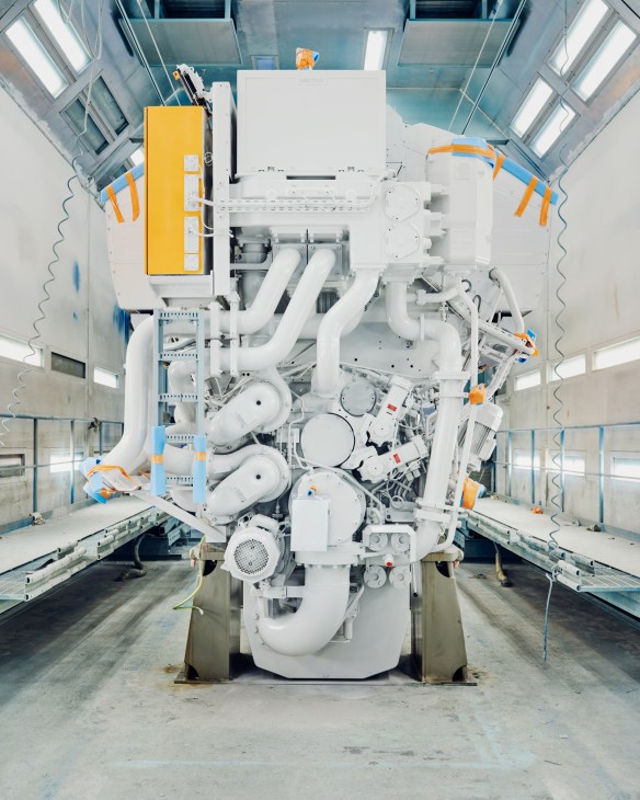 Overhead view of a large white marine engine with orange and yellow safety equipment in an industrial paint facility.