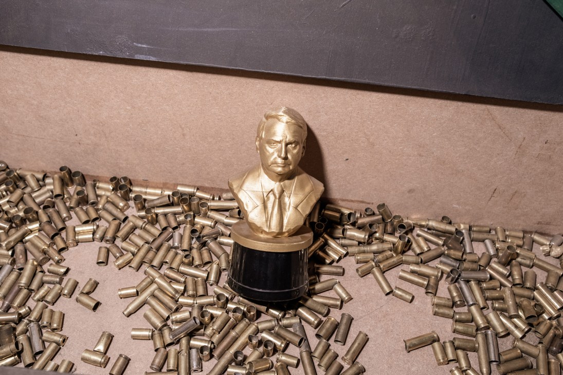 Golden bust of Jair Bolsonaro on black base surrounded by scattered bullet casings on concrete floor