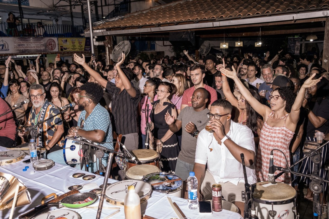 Crowded samba night with musicians playing percussion instruments while audience dances with raised hands
