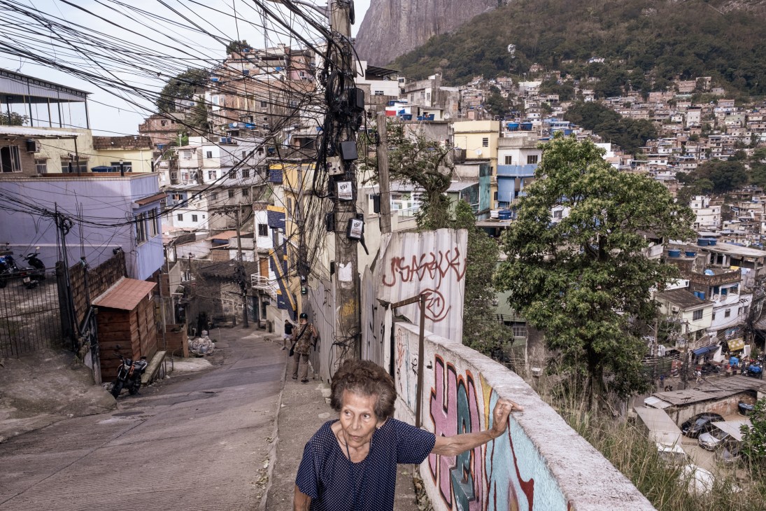 Elderly woman standing on concrete walkway overlooking densely packed favela buildings on hillside