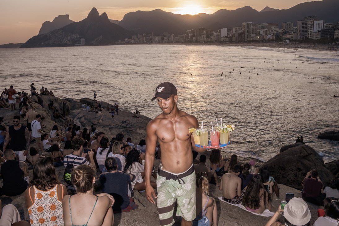 Shirtless man carrying colorful caipirinha cocktails on tray at crowded beach during sunset