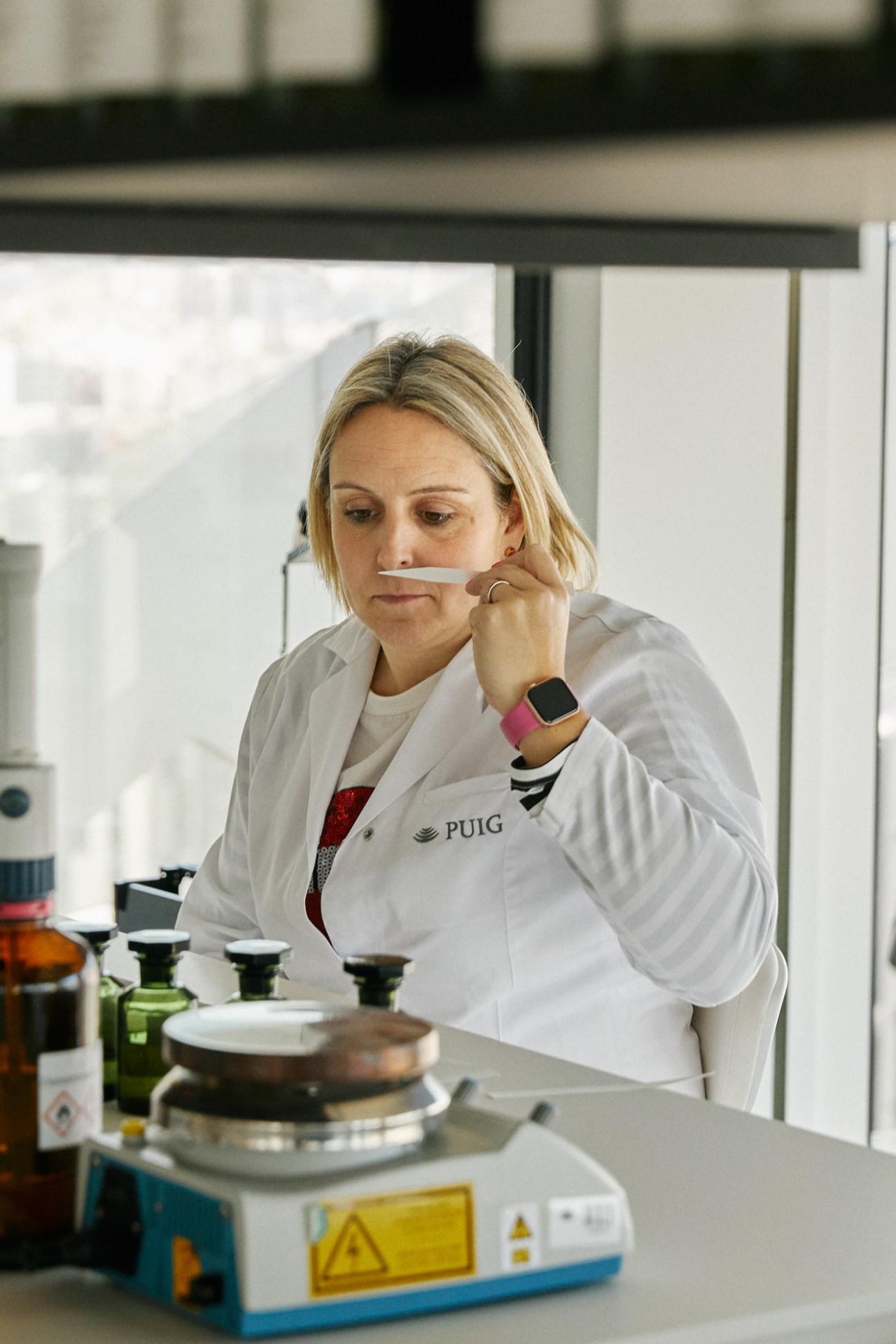 A woman in a white lab coat with 'PUIG' logo testing fragrances in a modern laboratory setting