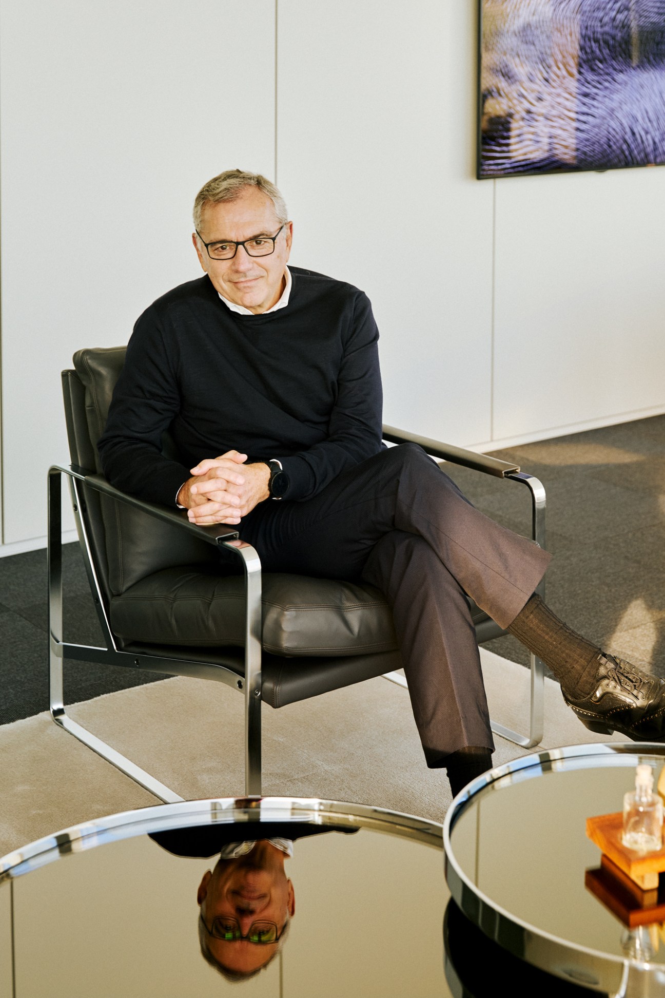 Marc Puig sits in a black leather chair wearing glasses, black sweater and dark trousers in a modern office