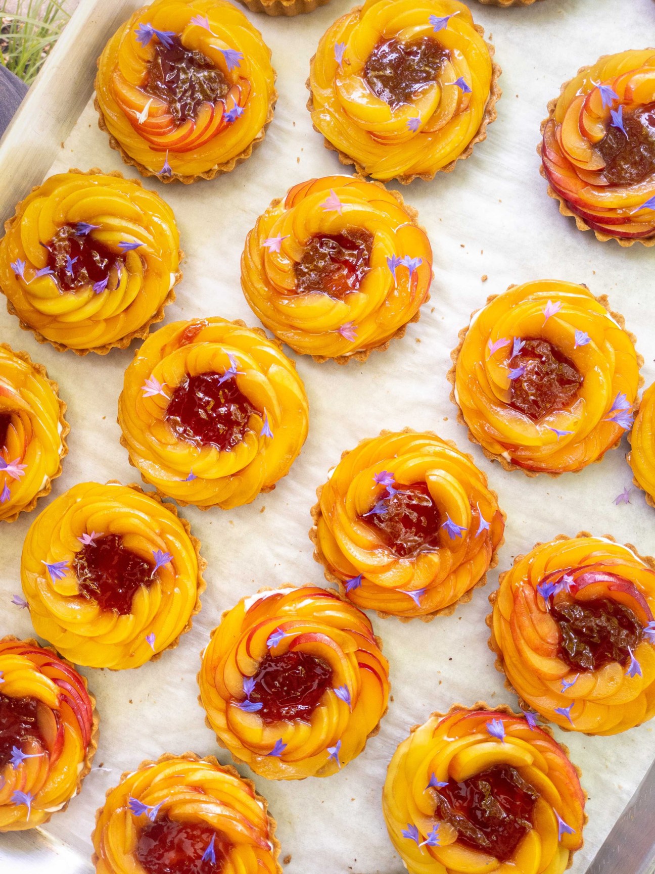 Golden pastry tarts with dark jam centers and purple flowers arranged on parchment paper