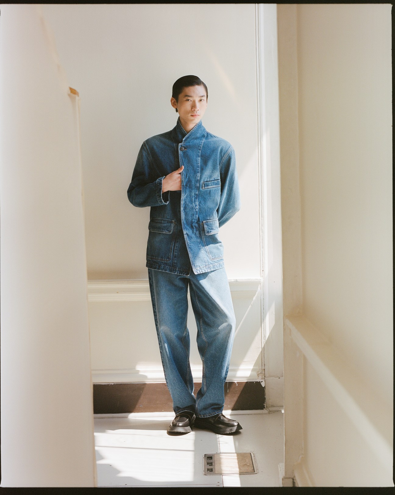 Model wearing Qasimi denim jacket and jeans in a minimalist white hallway.