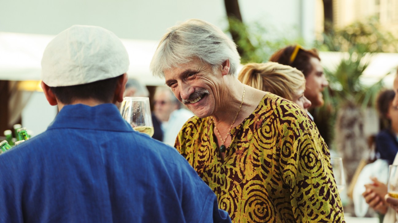 An older man with gray hair and mustache in a patterned shirt smiling while holding a wine glass at a social event