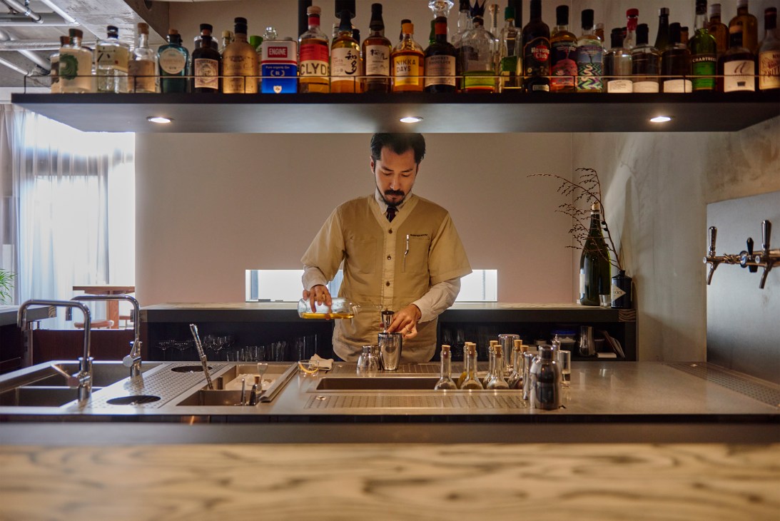 Bartender in tan vest preparing cocktails at Quarter Room bar with extensive bottle collection on overhead shelf