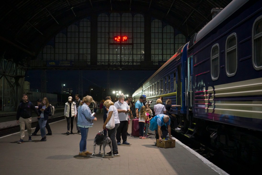 Passengers with luggage boarding a blue night train at a dimly lit railway station platform in the evening