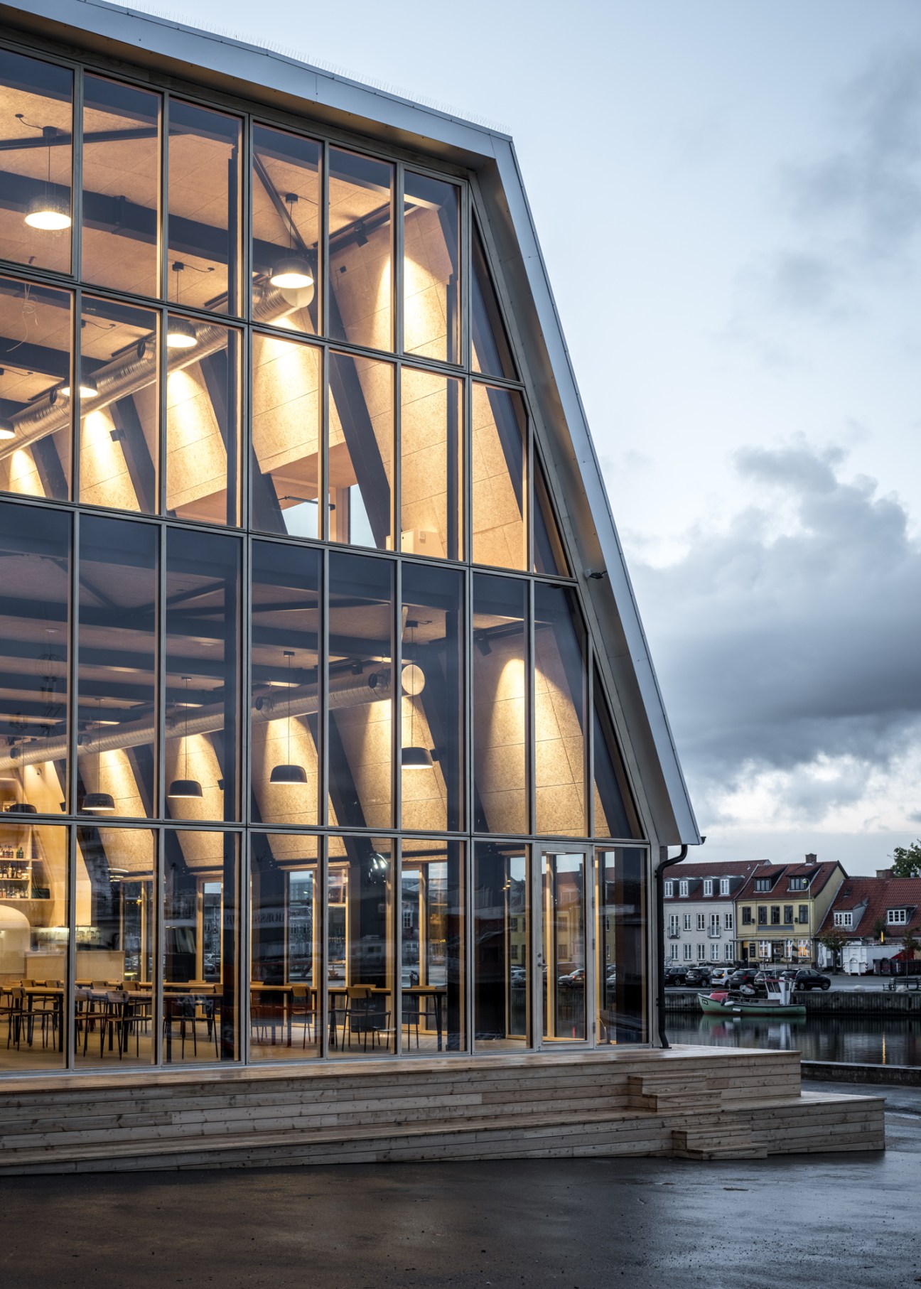 Modern glass building with exposed timber structure illuminated at dusk, featuring large windows and outdoor seating area