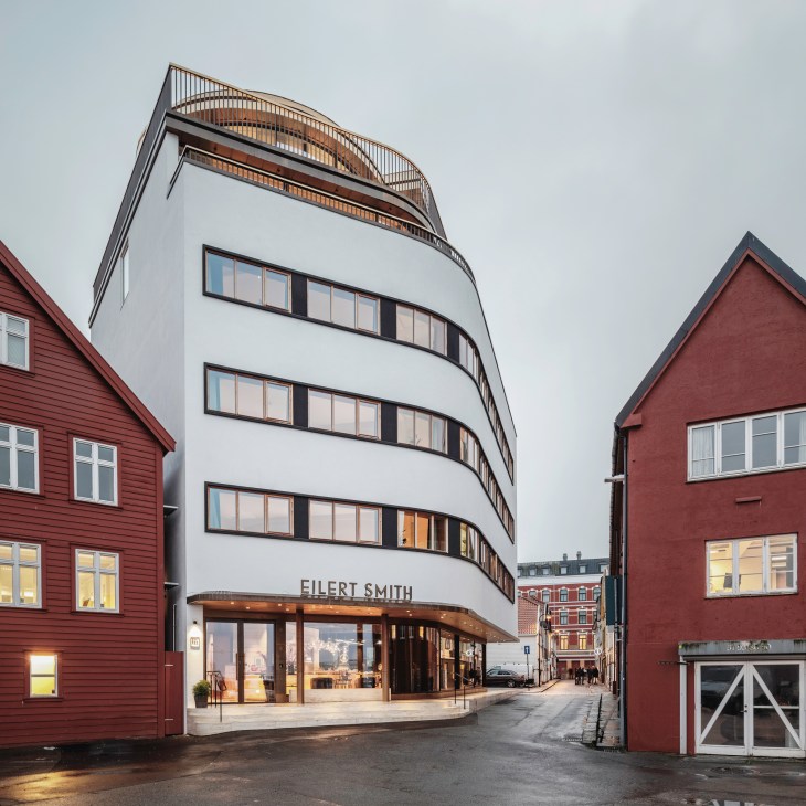 Modern white curved hotel building with horizontal ribbon windows nestled between traditional red wooden buildings