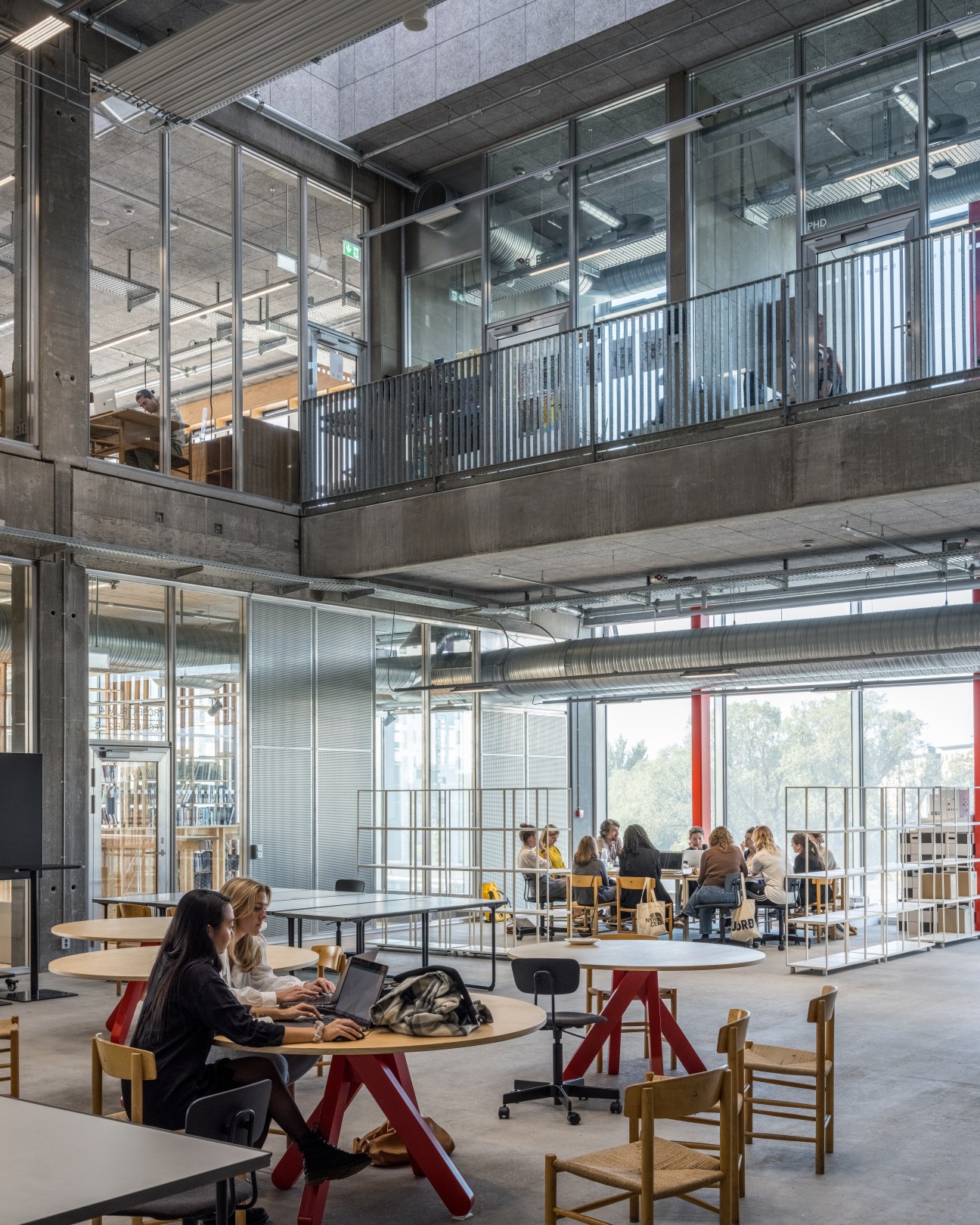 Modern open-plan workspace with mezzanine level, people working at tables with red legs, glass partitions, and industrial ceiling with skylights