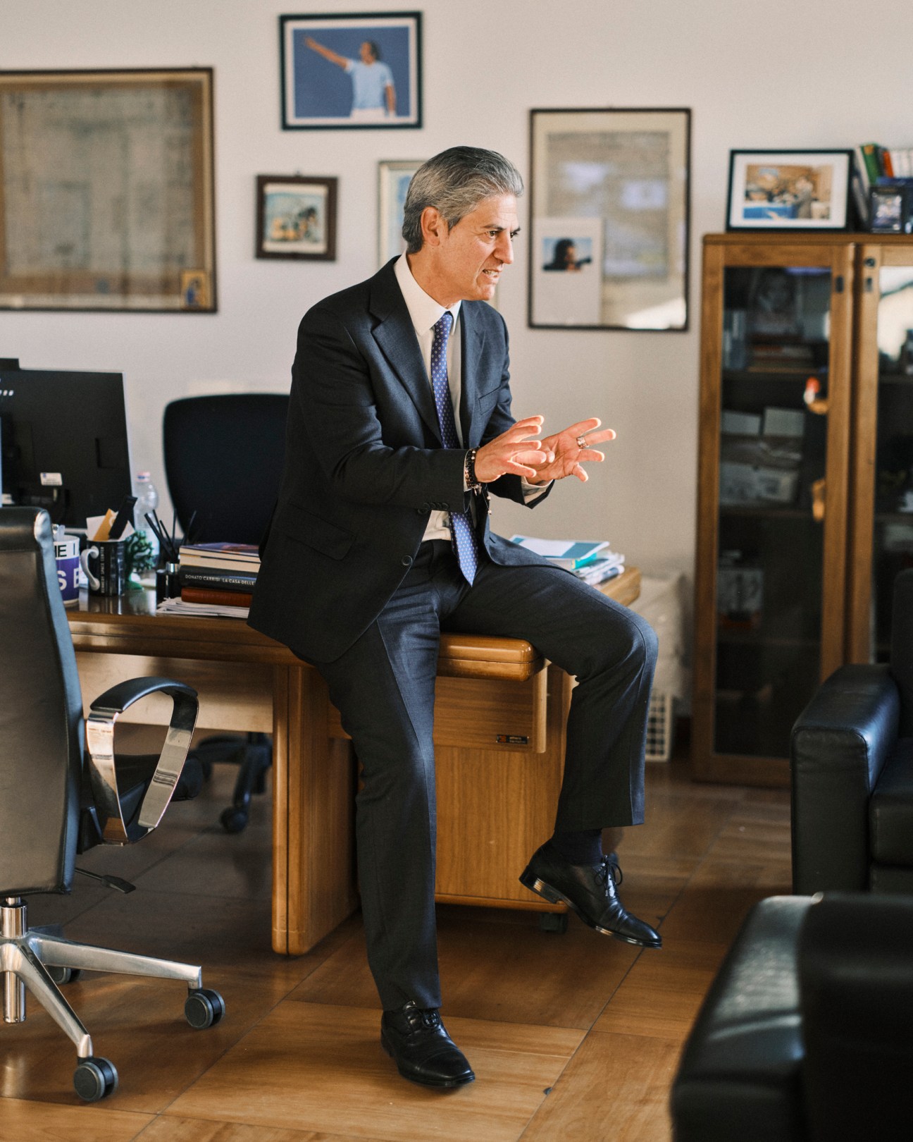Paolo Petrecca sits on a wooden desk in his office, gesturing with his hands while speaking
