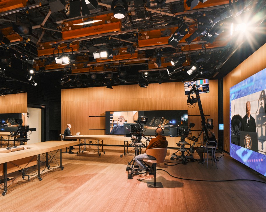 Television studio with cameras, lighting grid, wood desks, large screens and camera operator during live broadcast