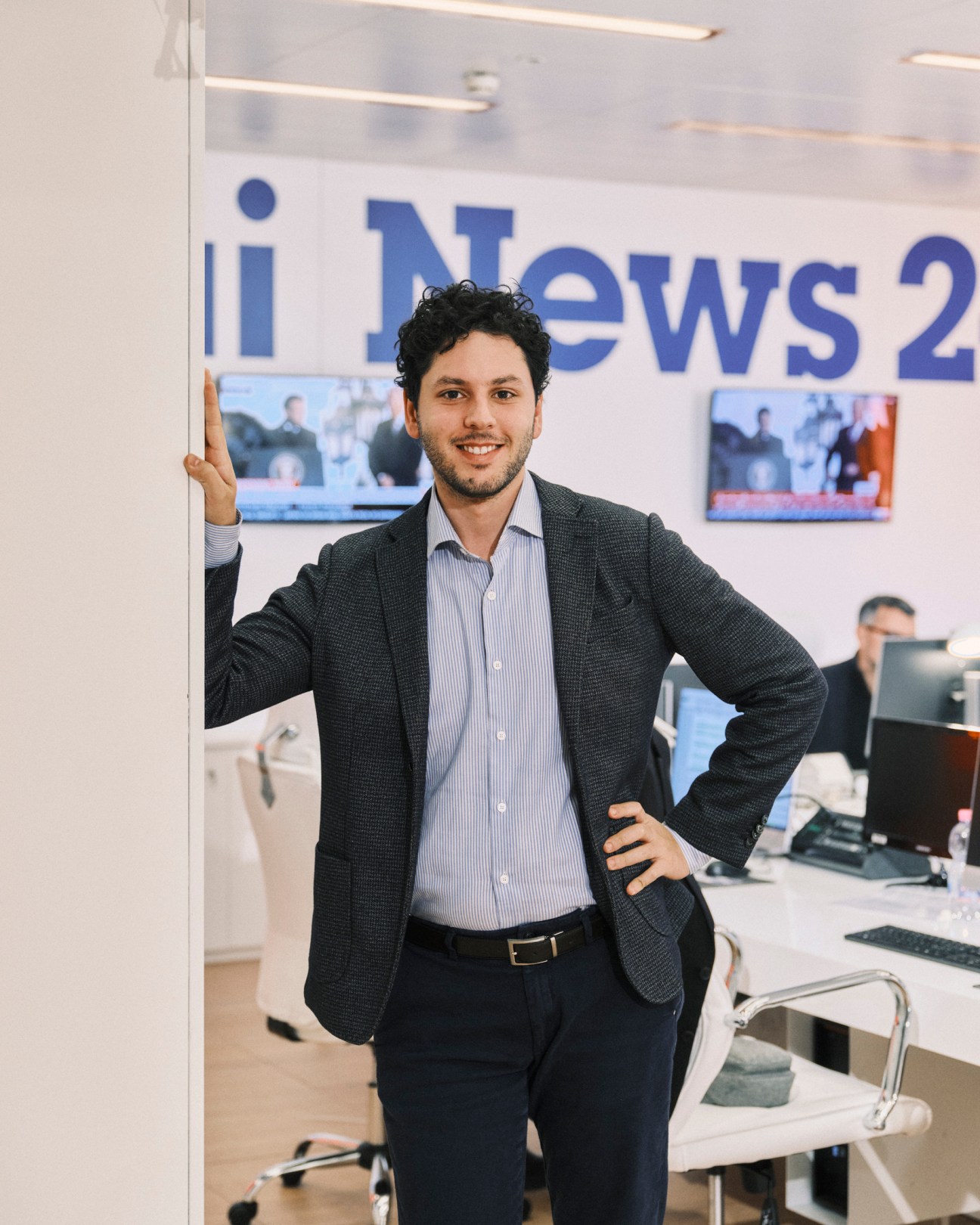Presenter Dario Marchetti standing in a modern TV newsroom with his hand on his hip