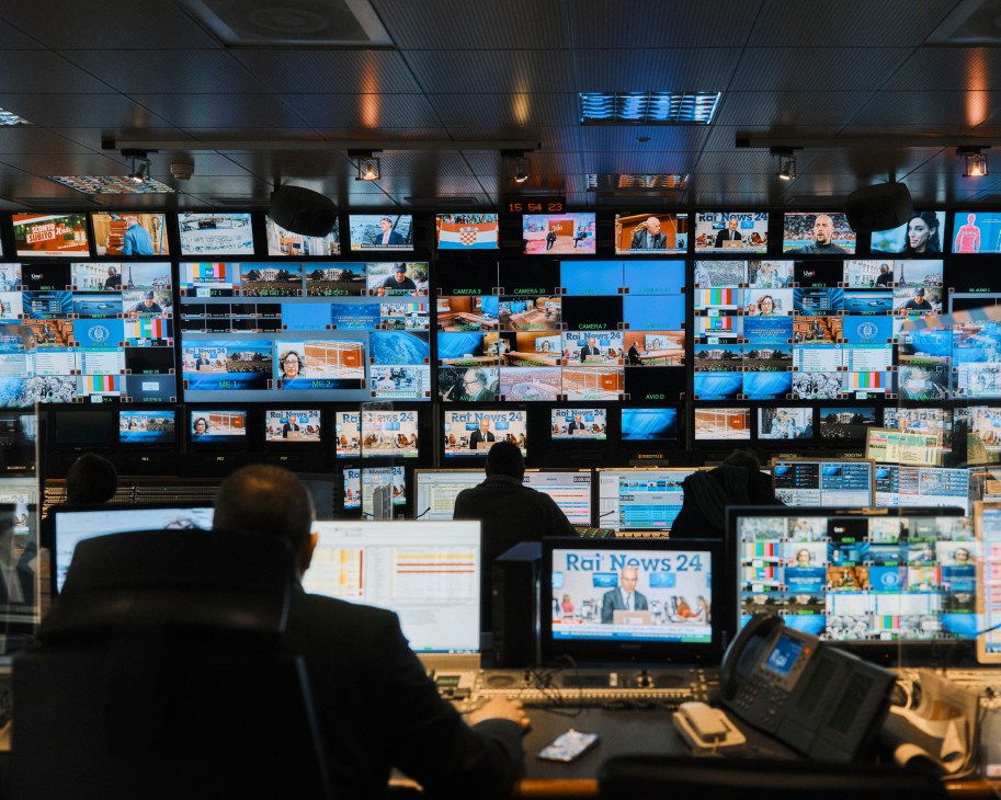 Control room with multiple monitors showing news broadcasts and silhouettes of operators at workstations