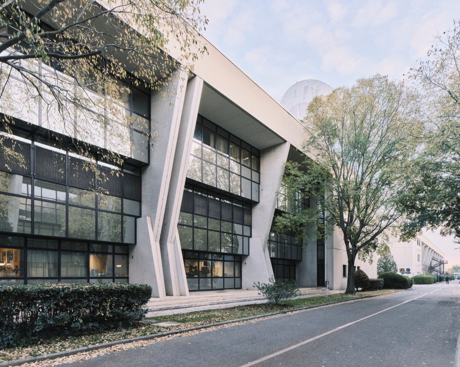Modern concrete building with angular white supports and large glass windows surrounded by trees