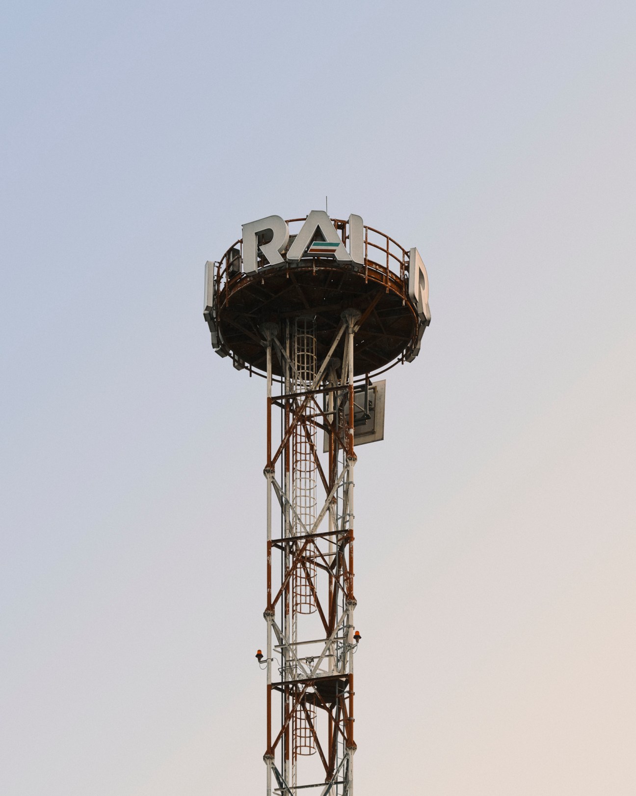Tall metal lattice transmission tower with RAI lettering on circular platform against pale sky