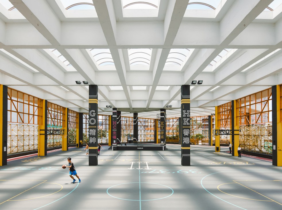 Modern basketball court with white ceiling beams, skylights, yellow structural elements and a person dribbling