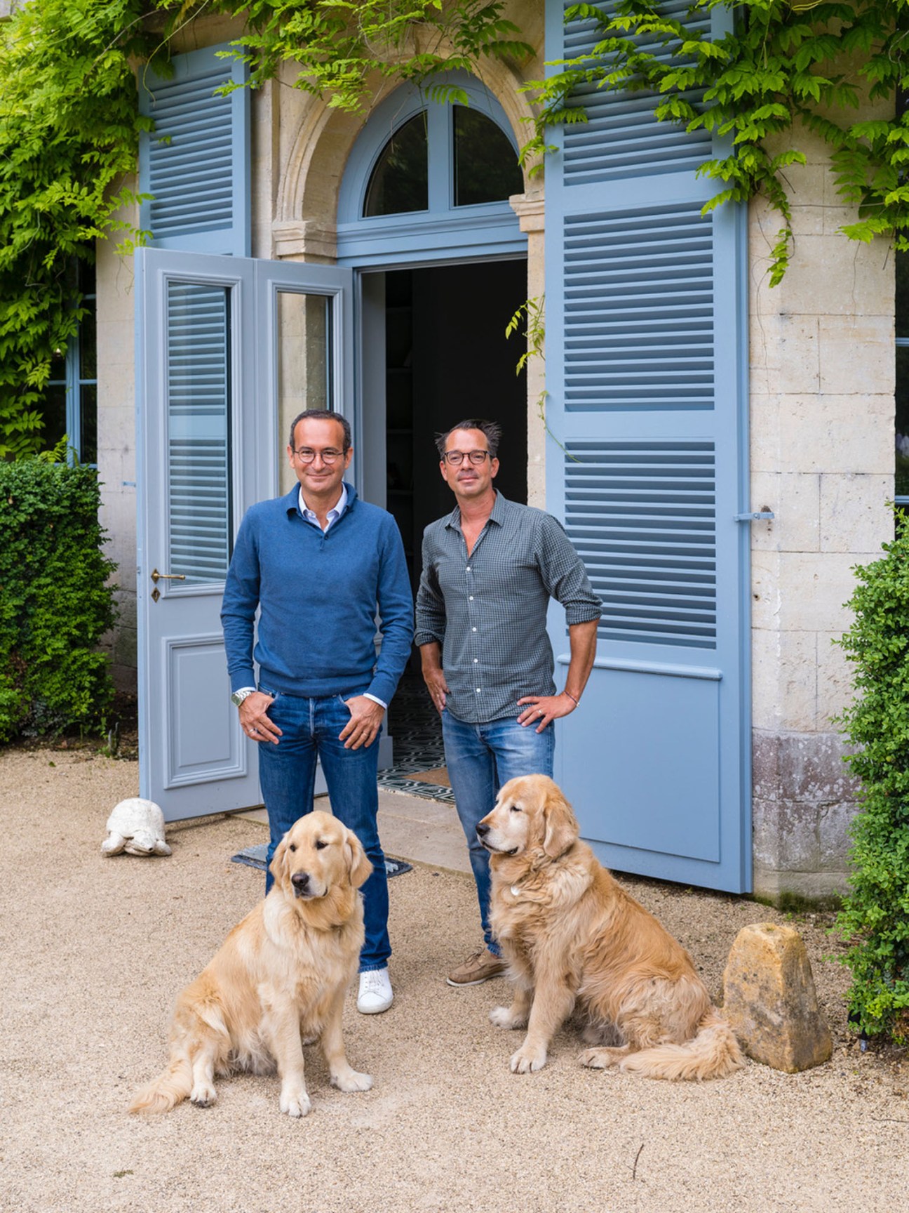 Two men standing with two golden retriever dogs in front of elegant French doors with blue shutters