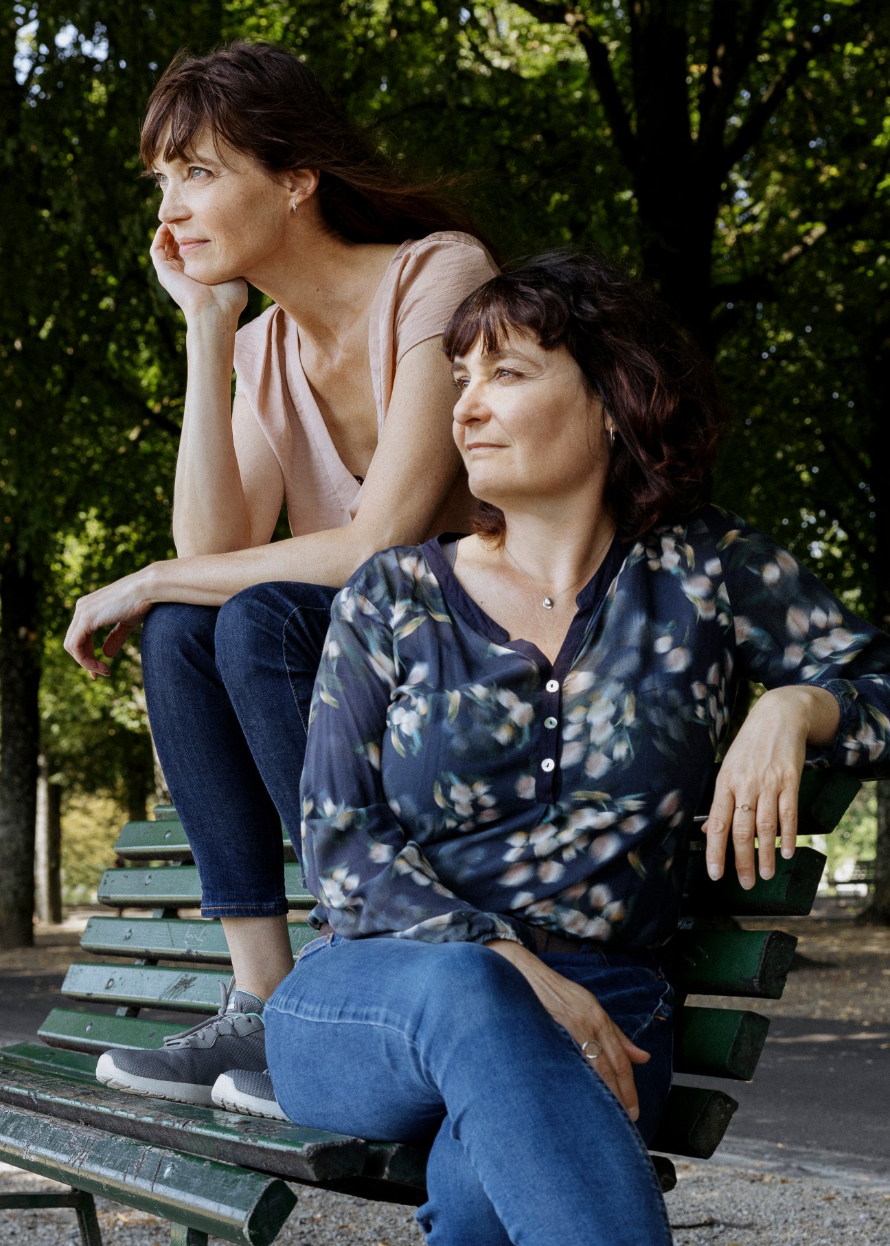 Two women sitting on a park bench in natural lighting, one behind the other