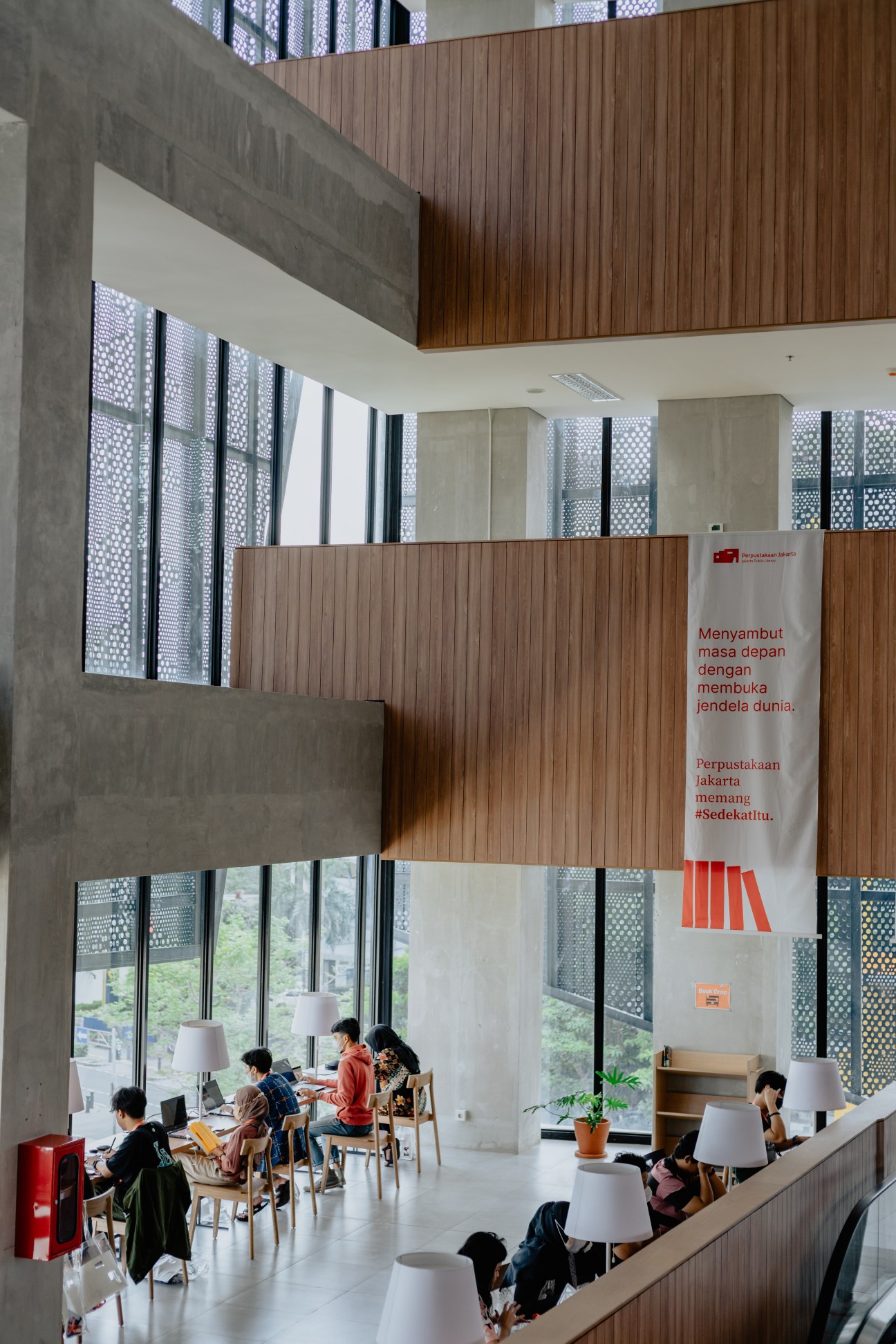 Interior of Jakarta's public library showing multi-level space with wooden ceiling, concrete walls, and visitors reading at tables.