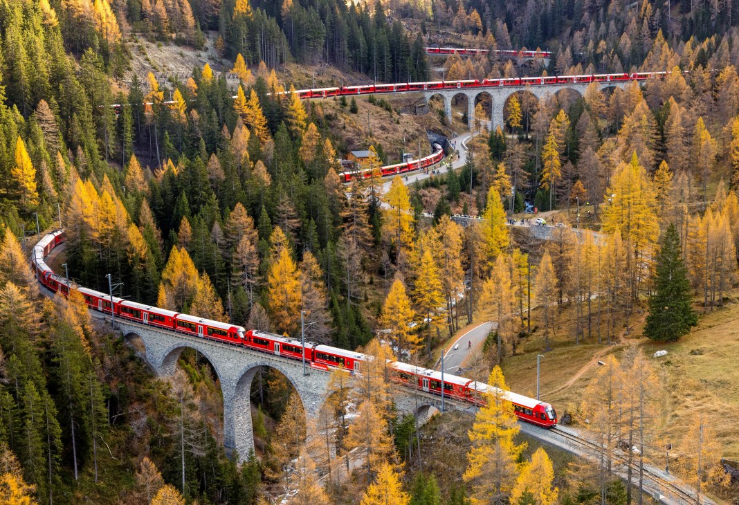 Red Rhätische Bahn train crossing stone viaduct through autumn forest in Swiss mountains
