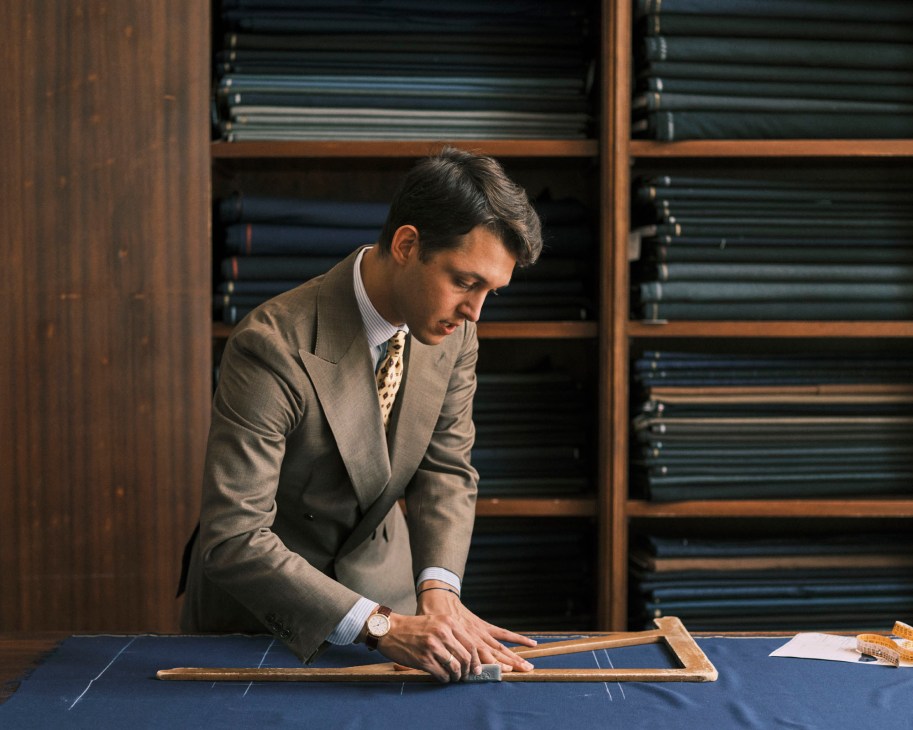 Young tailor in beige suit marking fabric with chalk on cutting table in workshop with fabric shelves