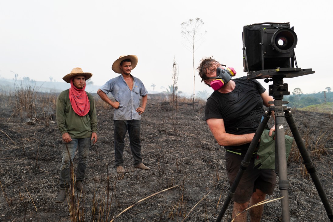 Photographer Richard Mosse operating a large camera on tripod in burned landscape with two local men watching