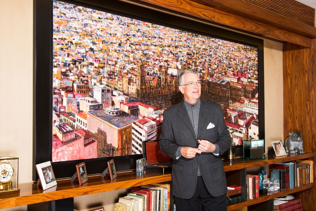 Ricardo Salinas stands in his office at TV Azteca in Mexico City, with a colorful cityscape artwork displayed on the wall behind him.