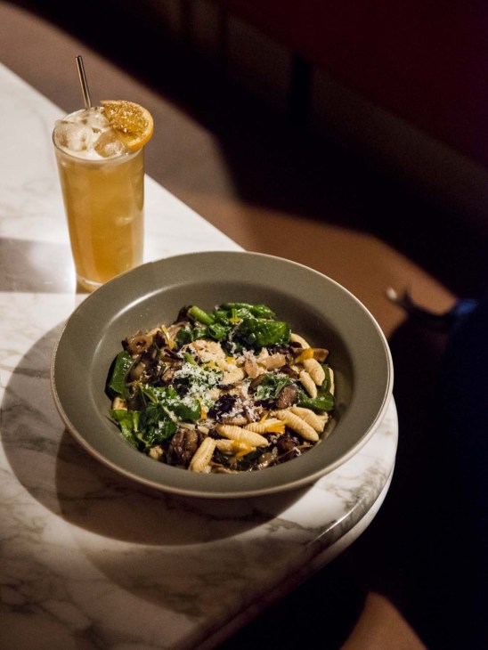 Ricotta cavatelli pasta dish with greens and grated cheese, served with a golden cocktail garnished with candied lemon.