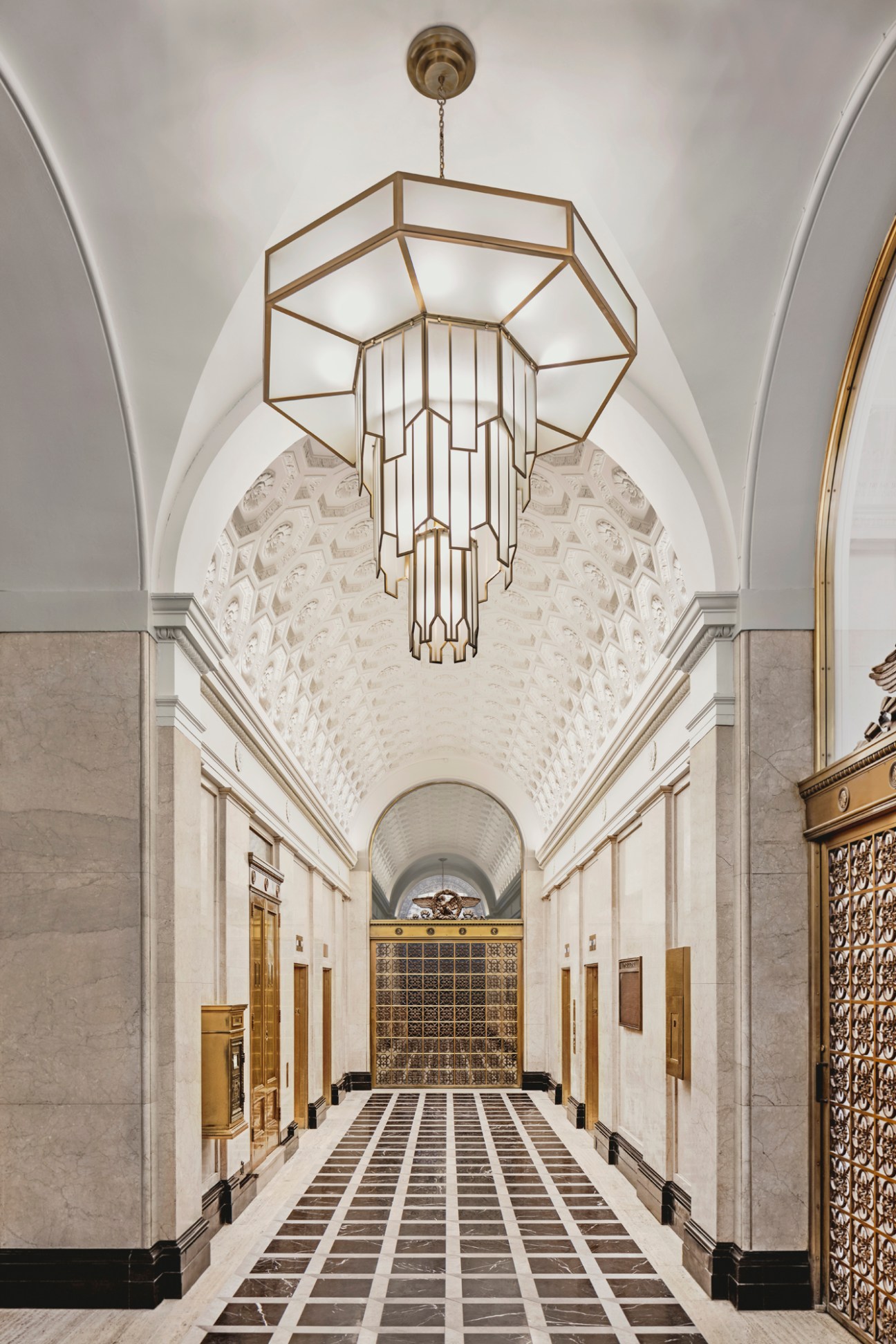 Grand marble corridor with geometric ceiling design and brass geometric chandelier hanging from center ceiling.