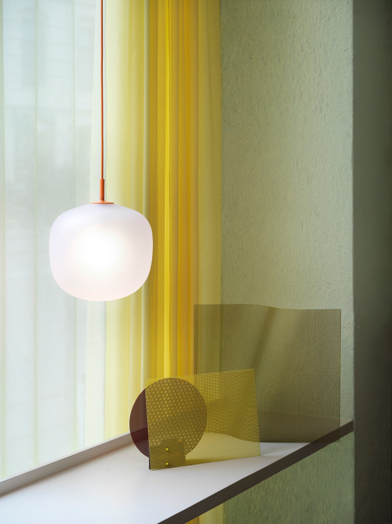 Rime Pendant Lamp by Muuto with mouth-blown glass globe and orange aluminum support pipe hanging near yellow curtains.