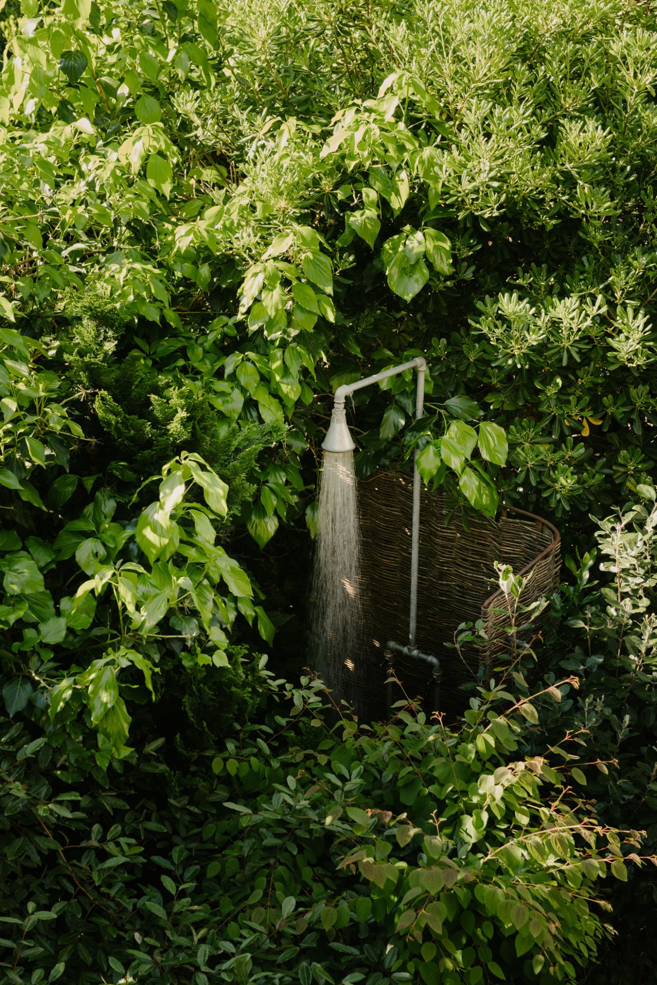 Outdoor shower with woven basket surrounded by lush green foliage and plants in a garden setting