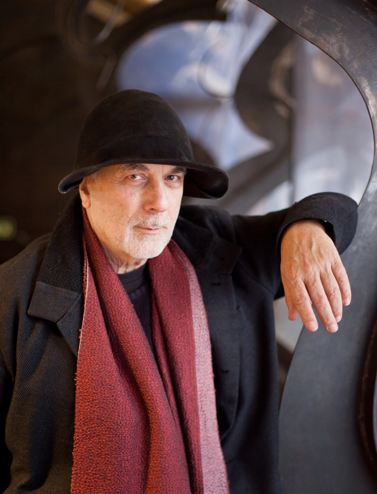 Ron Arad wearing black hat and red scarf, standing next to curved sculptural furniture piece in his design studio