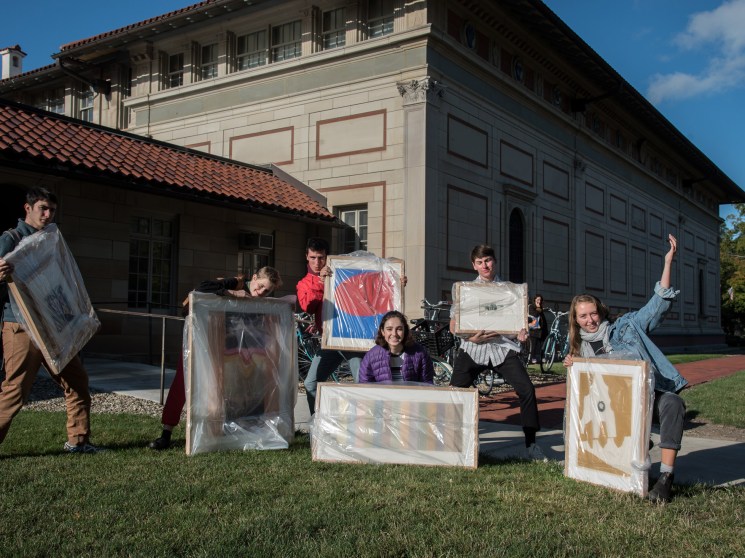 A group of students cheerfully display framed artworks on the lawn outside a classical stone building.