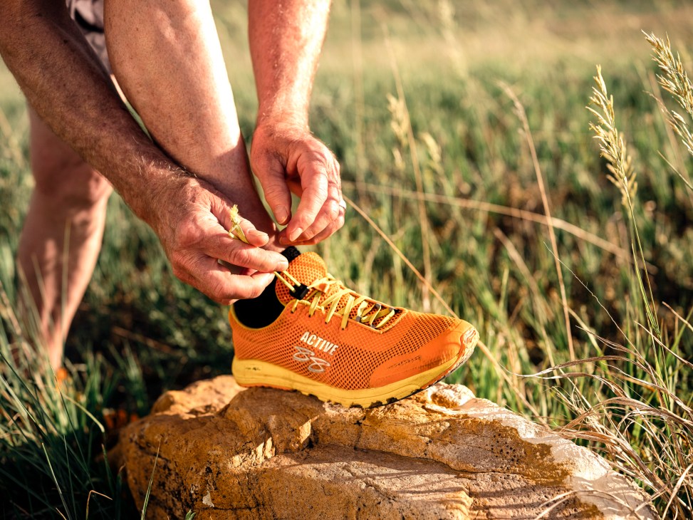 Hands lacing up an orange and yellow Active 88 running shoe on a rock in a grassy field.
