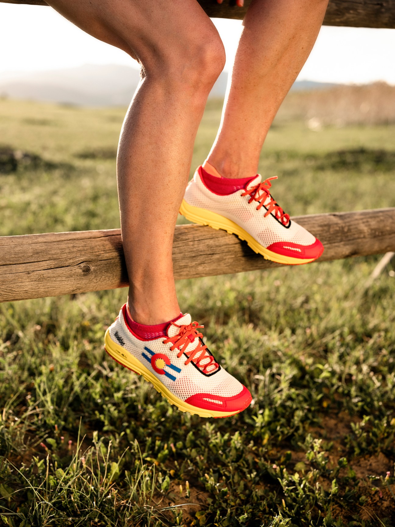 Person wearing Active 88 Boulder running shoes with red, white and yellow colorway on a wooden rail overlooking grassland.