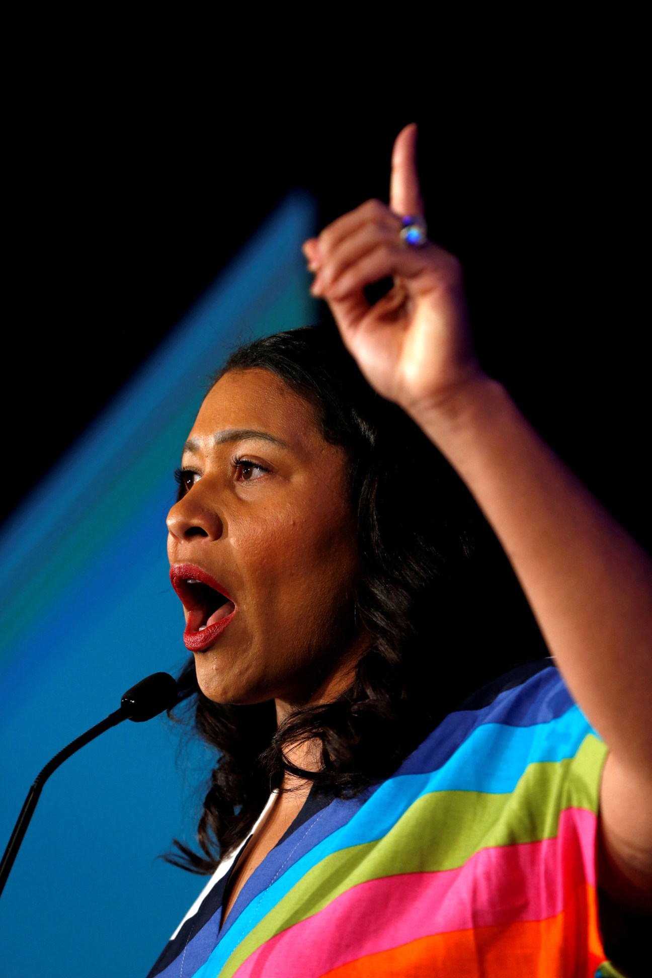 Woman speaking passionately at microphone with raised finger, wearing colorful striped blazer