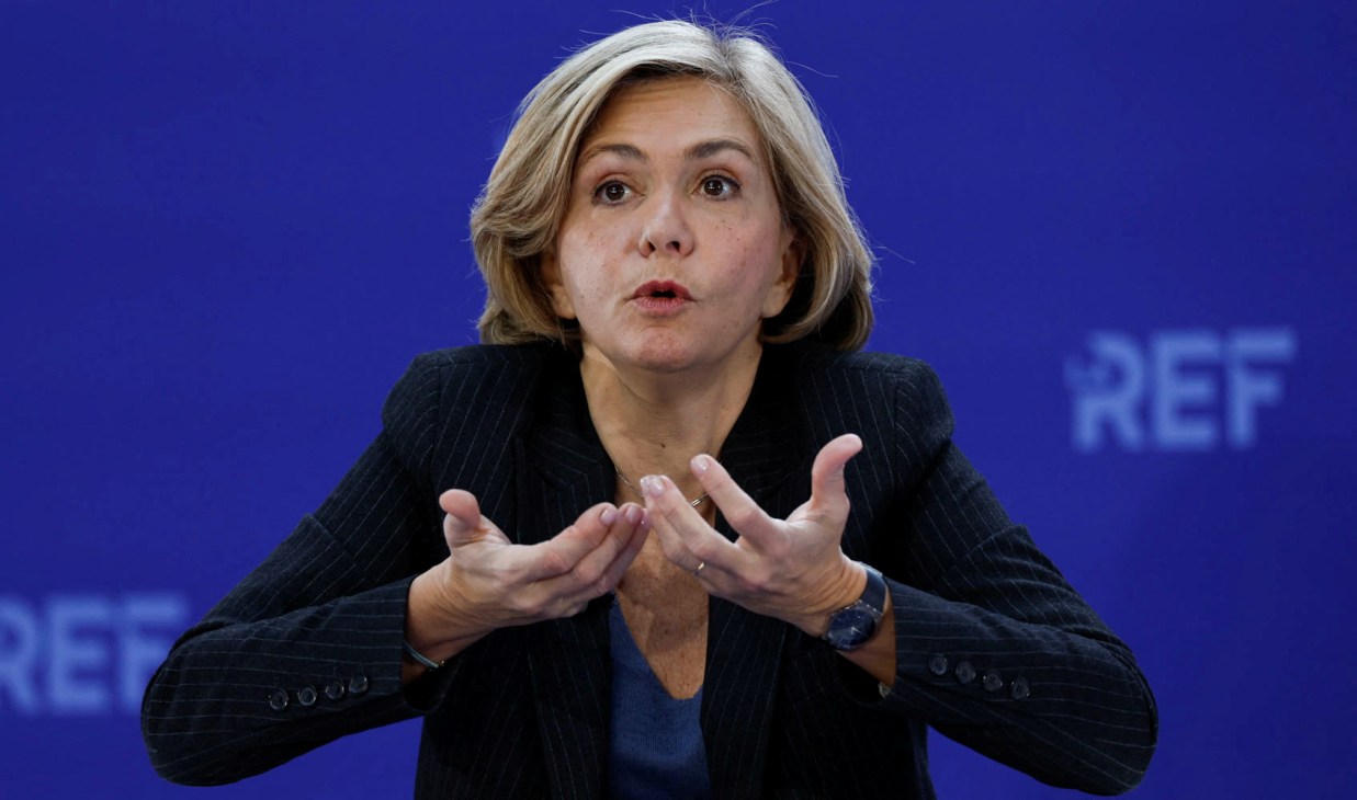 Woman in dark suit jacket gesturing with both hands while speaking at podium with blue background