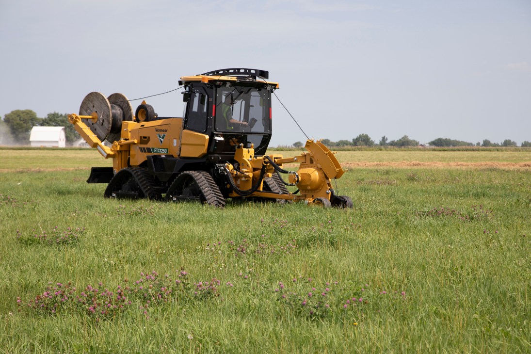 Yellow Vermeer RTX1250 tractor in a green field with wildflowers and farm buildings in background.
